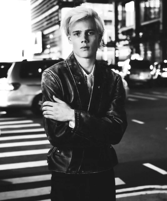 Black and white portrait of a young man in a leather jacket standing on a city street at night with buildings and traffic in background.