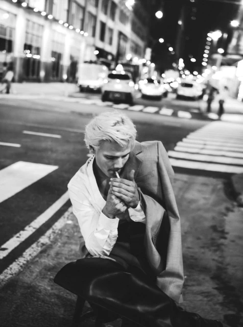 Monochrome portrait of a fair-skinned adult male sitting on an urban street at night, lighting a cigarette near city traffic.
