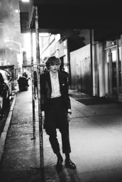 Young male model in dark suit jacket and trousers leaning against a pole on a city sidewalk at night