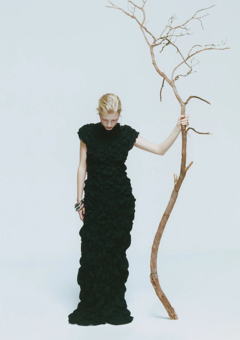 A woman in a long black dress poses in front of a white background holding a bare branch in a fashion editorial studio shot.