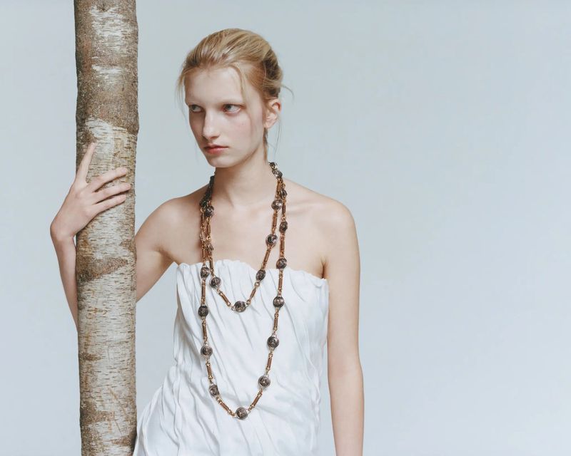 A fair-skinned woman with blond hair stands next to a birch tree, wearing a white strapless top and a long beaded necklace in a photo studio.