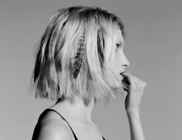Monochrome portrait of a woman in profile with short blonde hair, feathers, and dark roots against a light background.
