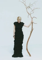 Model in a black textured dress holding a bare tree branch against a plain white studio background.