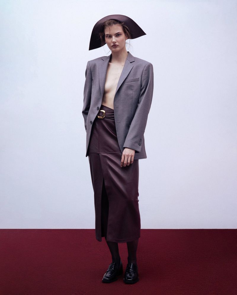 Model wearing an oversized grey blazer, brown leather skirt, and wide-brimmed hat against a plain studio background.