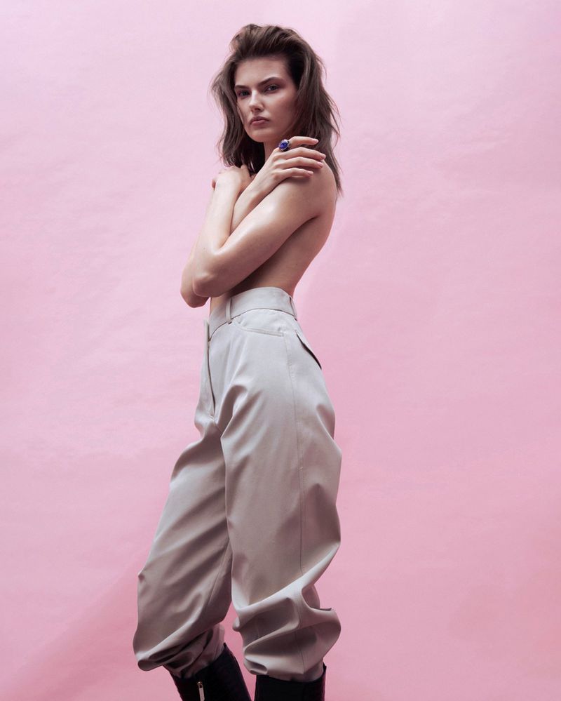 Model wearing high-waisted grey trousers and dark boots against a solid pink background for Harper's Bazaar March 2026.