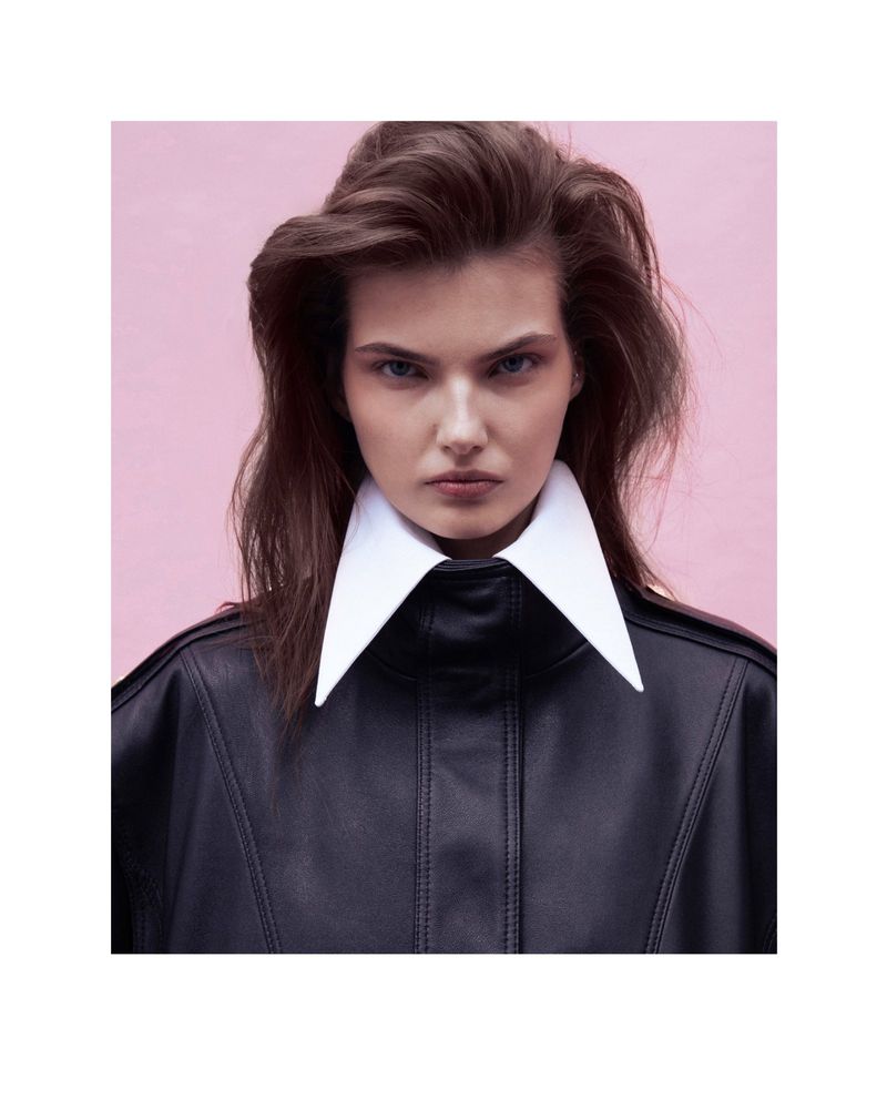 Brunette woman with a neutral expression wears a black jacket with a white collar in a medium studio shot against a pink backdrop.
