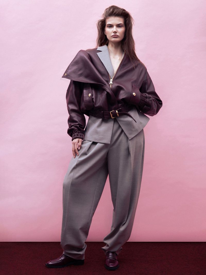 Fashion editorial shot of a female model in an oversized brown leather jacket and grey pants, standing against a pink background.