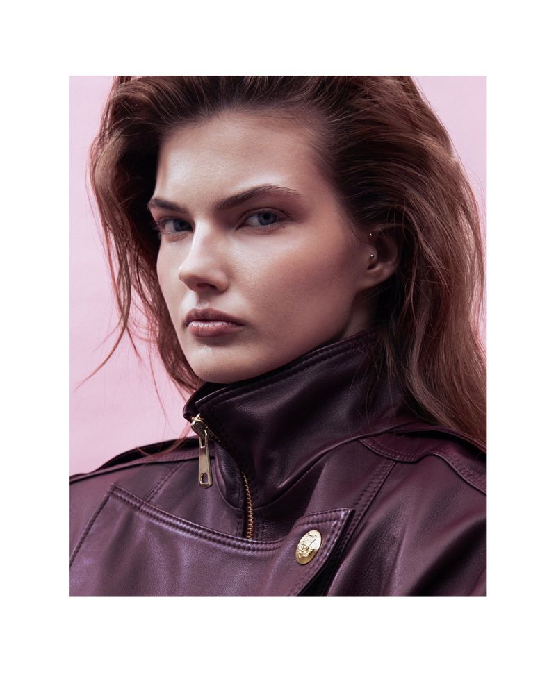 Portrait of a fair-skinned young woman with brown hair and neutral expression, wearing a brown leather jacket against a pink backdrop.