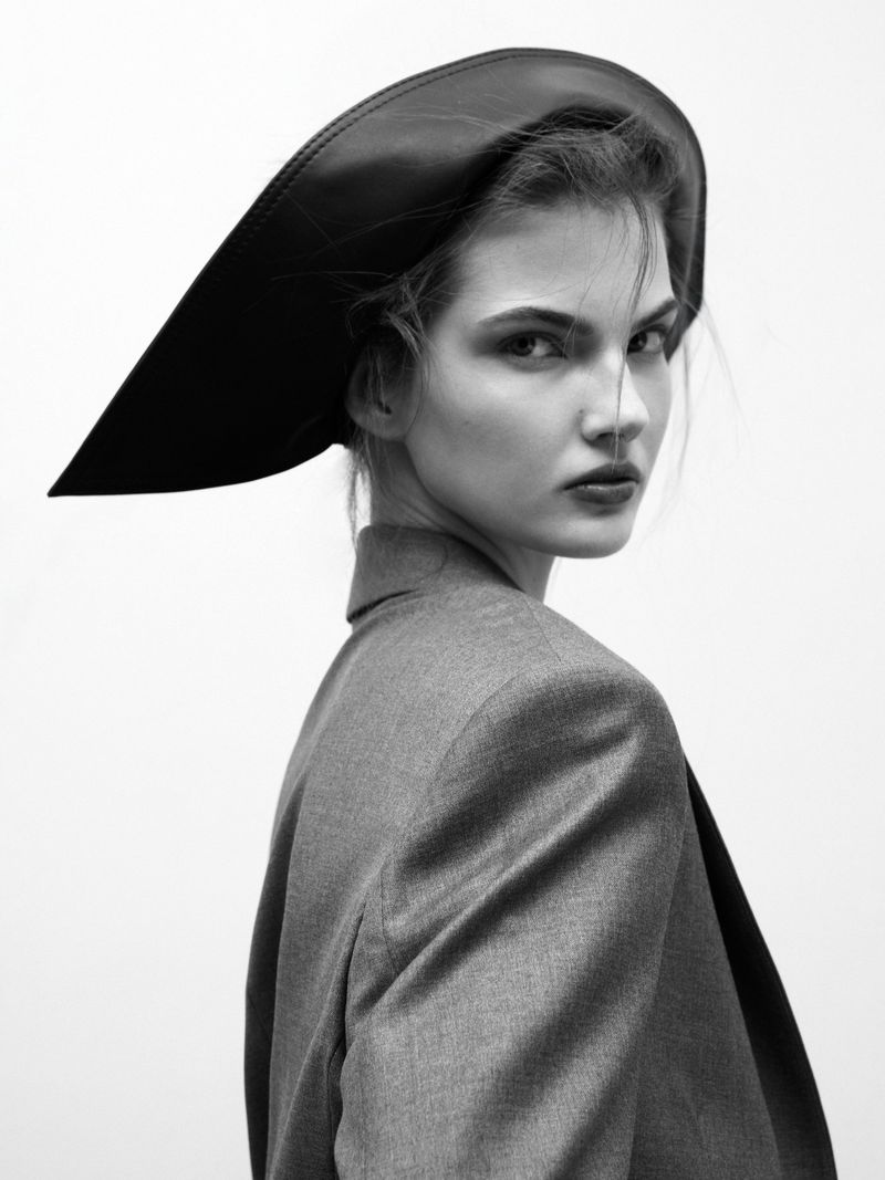 Monochrome portrait of an adult female fashion model in a blazer and large brim hat looking over her shoulder in a photo studio.
