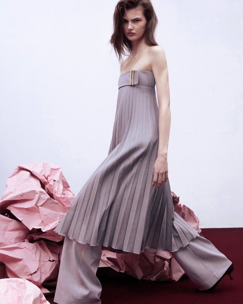 A young woman with shoulder-length hair poses in a strapless pleated top and wide-leg pants, standing on a red surface amidst crumpled pink paper.
