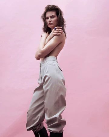 Model wearing high-waisted grey trousers and dark boots against a solid pink background for Harper's Bazaar March 2026.