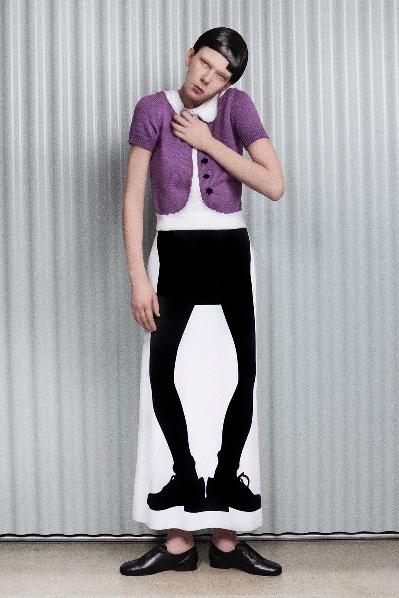 Woman in white dress with black leggings print, purple cardigan and black flats poses in a studio with a corrugated metal backdrop.