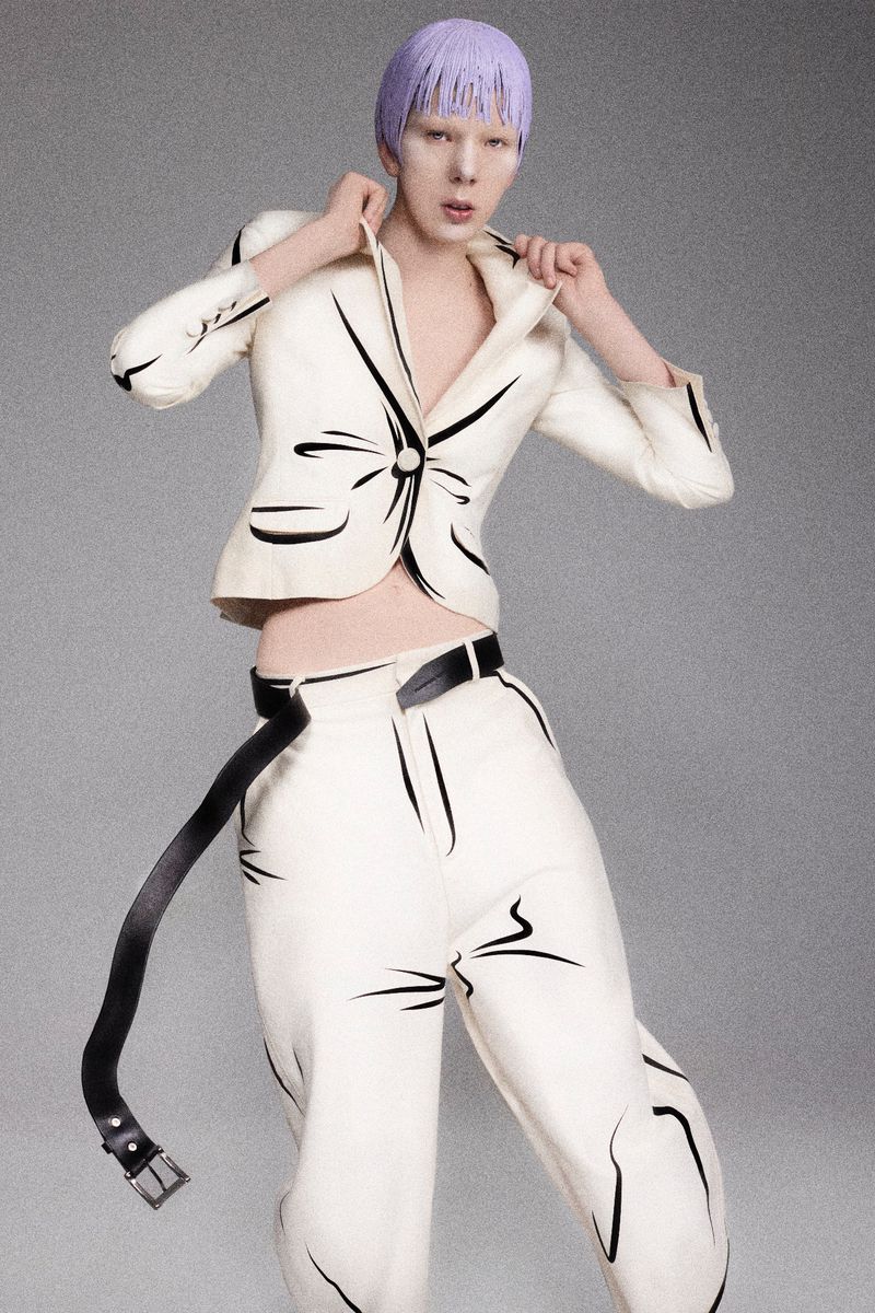 Fashion-forward adult posing indoors with white pantsuit with black line art details, black belt, and pastel purple hair in studio lighting.