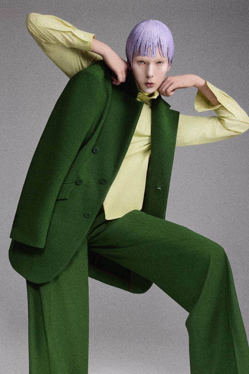 A fashion-forward model poses in a studio, wearing an oversized green pantsuit and a light yellow blouse, accented by a lavender hairstyle.