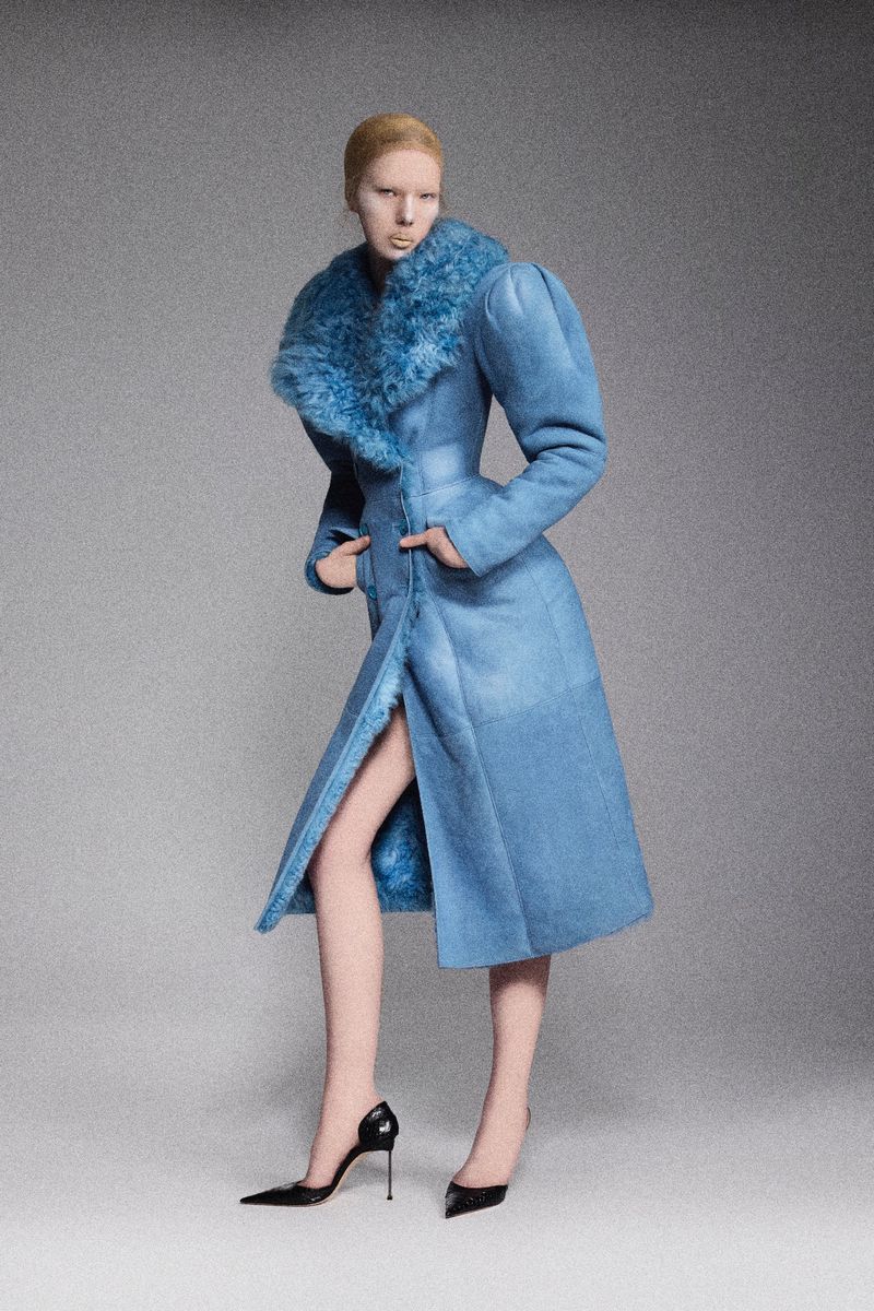 A blonde fashion model poses in a full-length light blue coat with a large faux fur collar, black patent leather high heels, and a grey background.