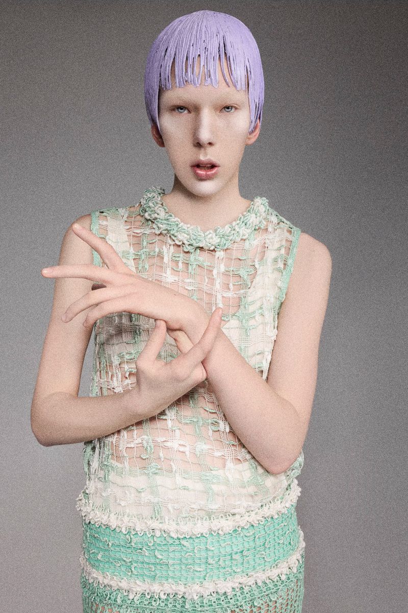 Close-up of a person with pale skin, lavender hair, and a light green and white woven garment in a studio setting, against a gray background.