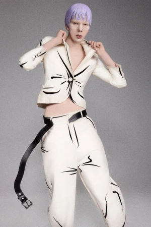 Model with lavender hair wearing cream jacket and trousers with black brushstroke details against a grey seamless background