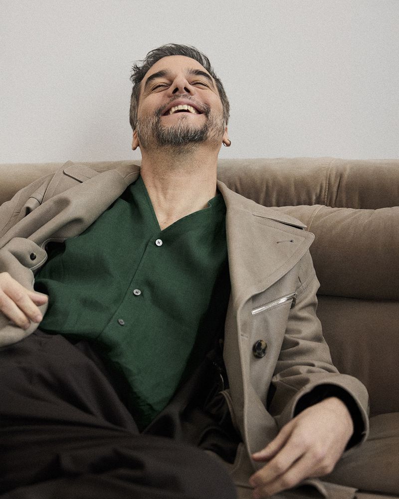 Middle-aged man with dark hair and beard laughs while lounging on a brown couch wearing a light-brown trench coat and dark-green button-down.