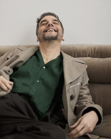 Middle-aged man with dark hair and beard laughs while lounging on a brown couch wearing a light-brown trench coat and dark-green button-down.