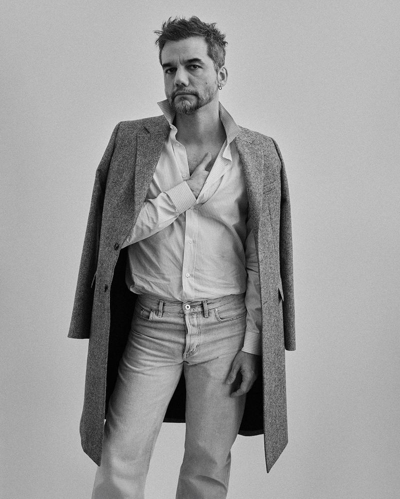 Black and white portrait of an adult man in an open collared shirt, light denim jeans, and a coat draped over his shoulders against white.