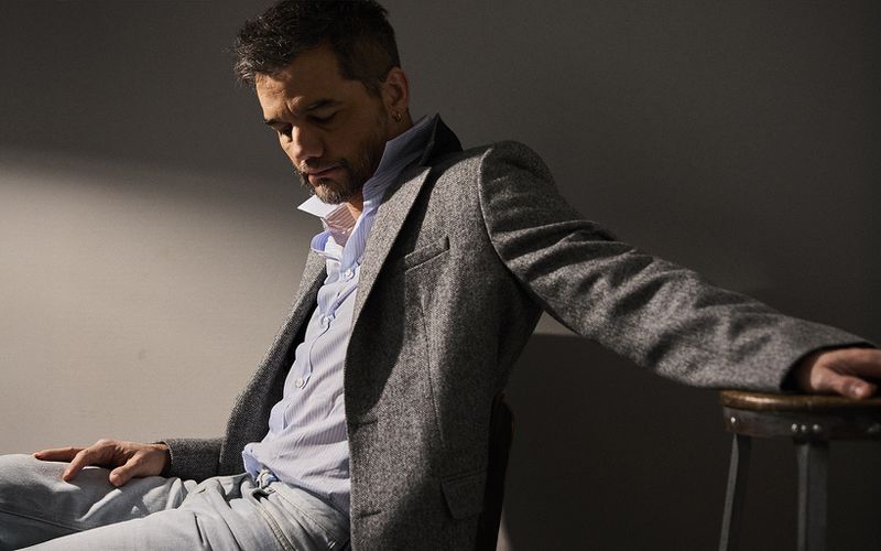 Man in a grey blazer and light blue jeans sits with a hand resting on a dark stool, looking down in a neutral-toned indoor setting.