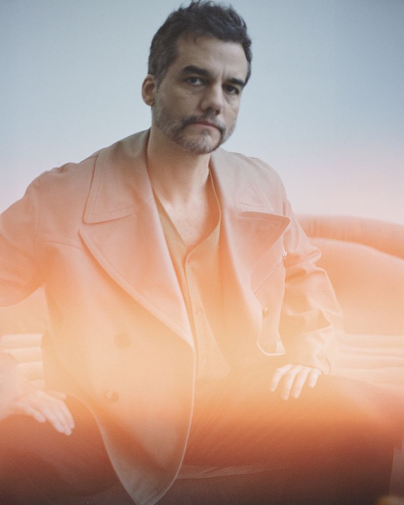 Portrait of adult man with stubble in neutral clothing, sitting on coral sofa with orange light leak and muted blue wall backdrop.