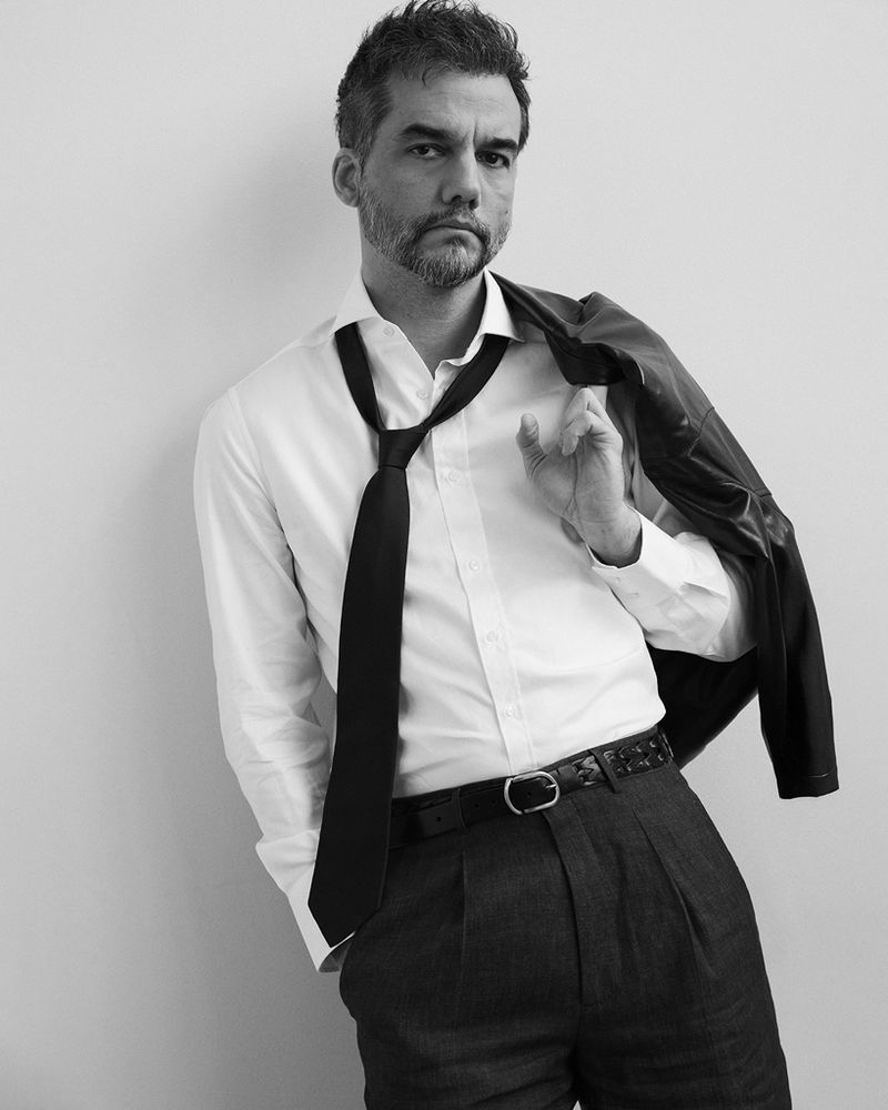Portrait of Wagner Moura wearing a white shirt, black tie and dark trousers, holding a jacket over his shoulder.