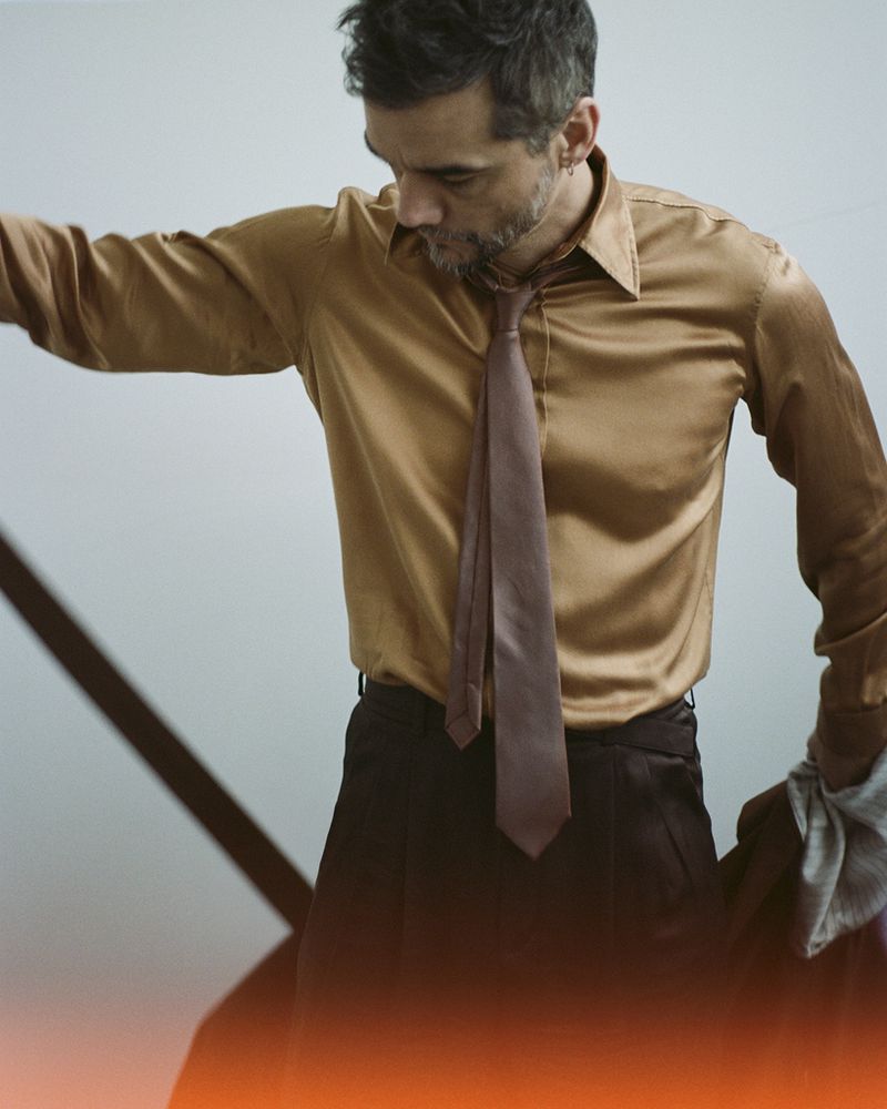 An adult male, casually posed in a gold shirt and tie, is indoors near a decorative black metal object with a neutral-toned background.