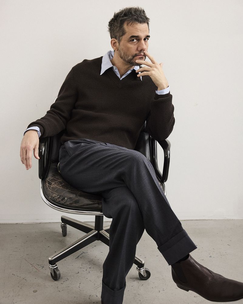 Man in brown sweater sitting in black leather office chair against a white wall, looking directly at the camera with a contemplative expression.
