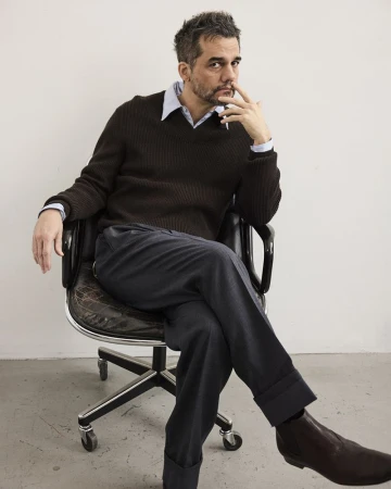 Man in brown sweater sitting in black leather office chair against a white wall, looking directly at the camera with a contemplative expression.