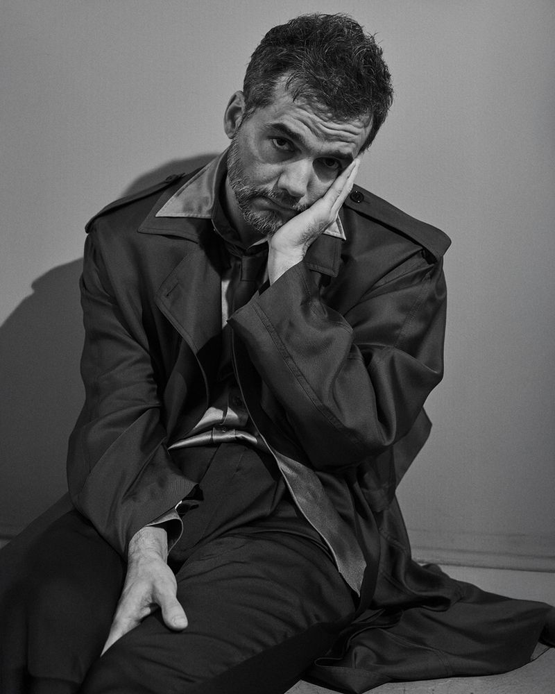 Portrait of a melancholic man with a beard, wearing a satin coat and pants, resting his face on his hand in a photo studio setting.