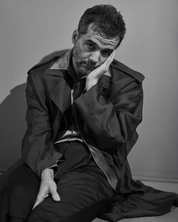 Portrait of a melancholic man with a beard, wearing a satin coat and pants, resting his face on his hand in a photo studio setting.