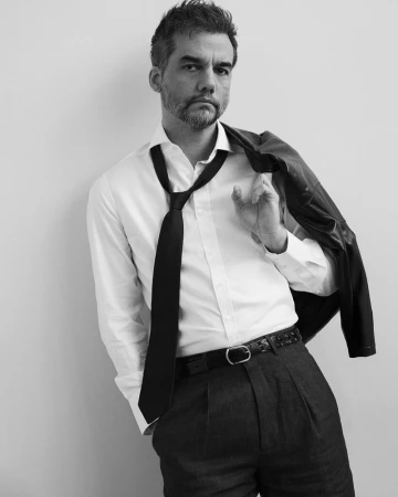 Portrait of Wagner Moura wearing a white shirt, black tie and dark trousers, holding a jacket over his shoulder.
