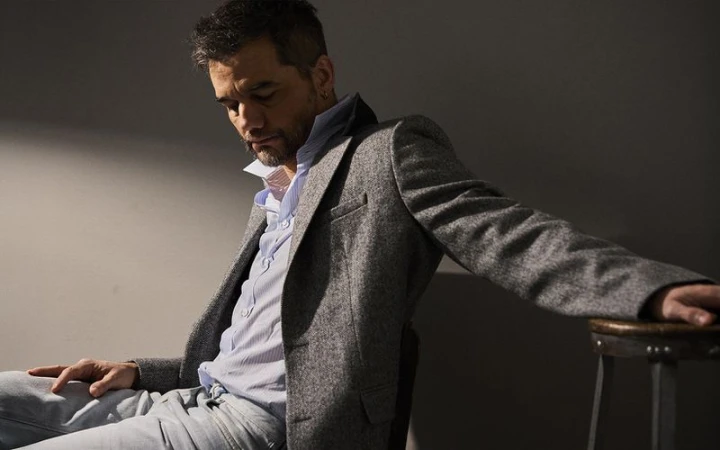 Man wearing a grey tweed blazer and light blue shirt seated against a plain dark wall in a studio portrait.