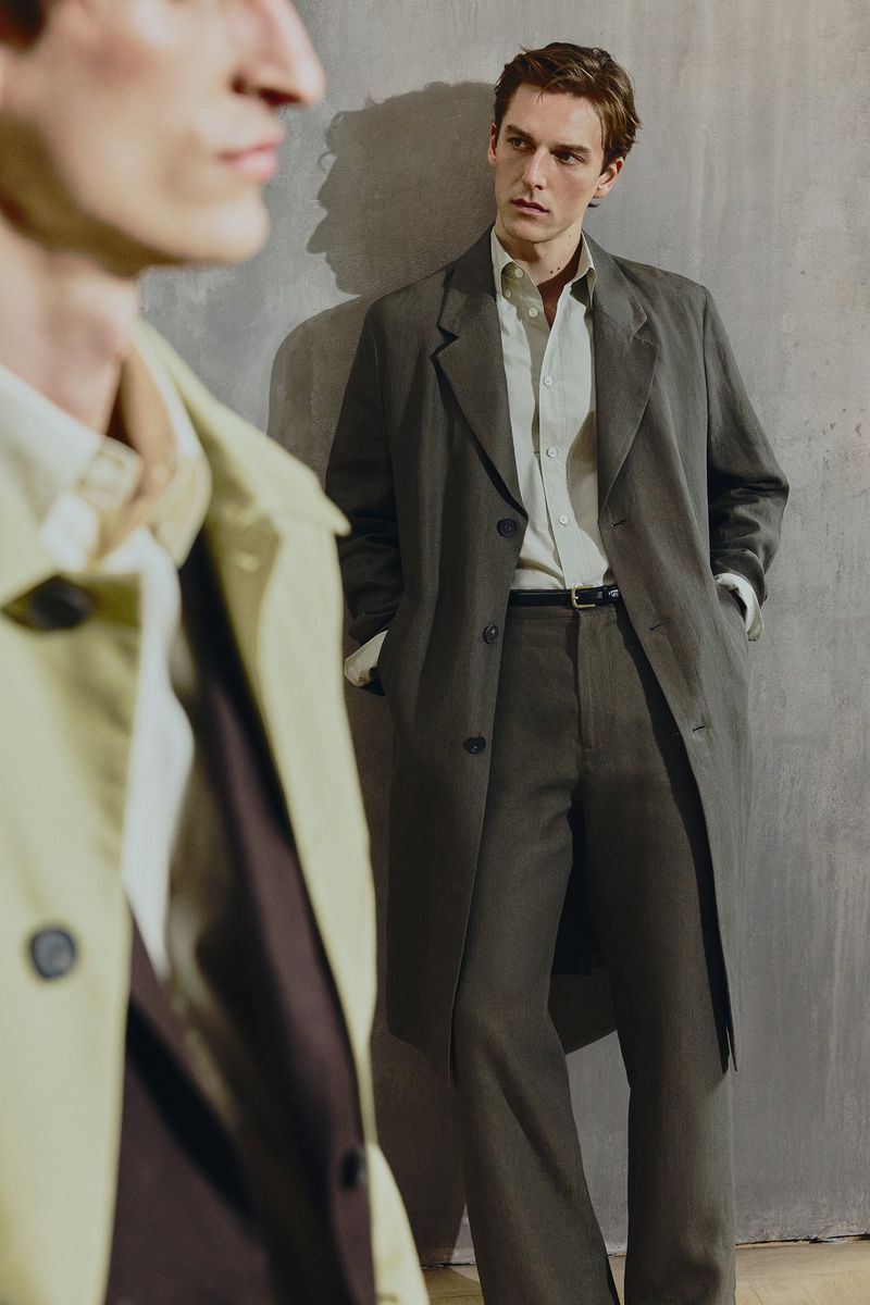 Two adult men in neutral-toned overcoats pose in a minimalist indoor setting, creating a sophisticated fashion editorial look.