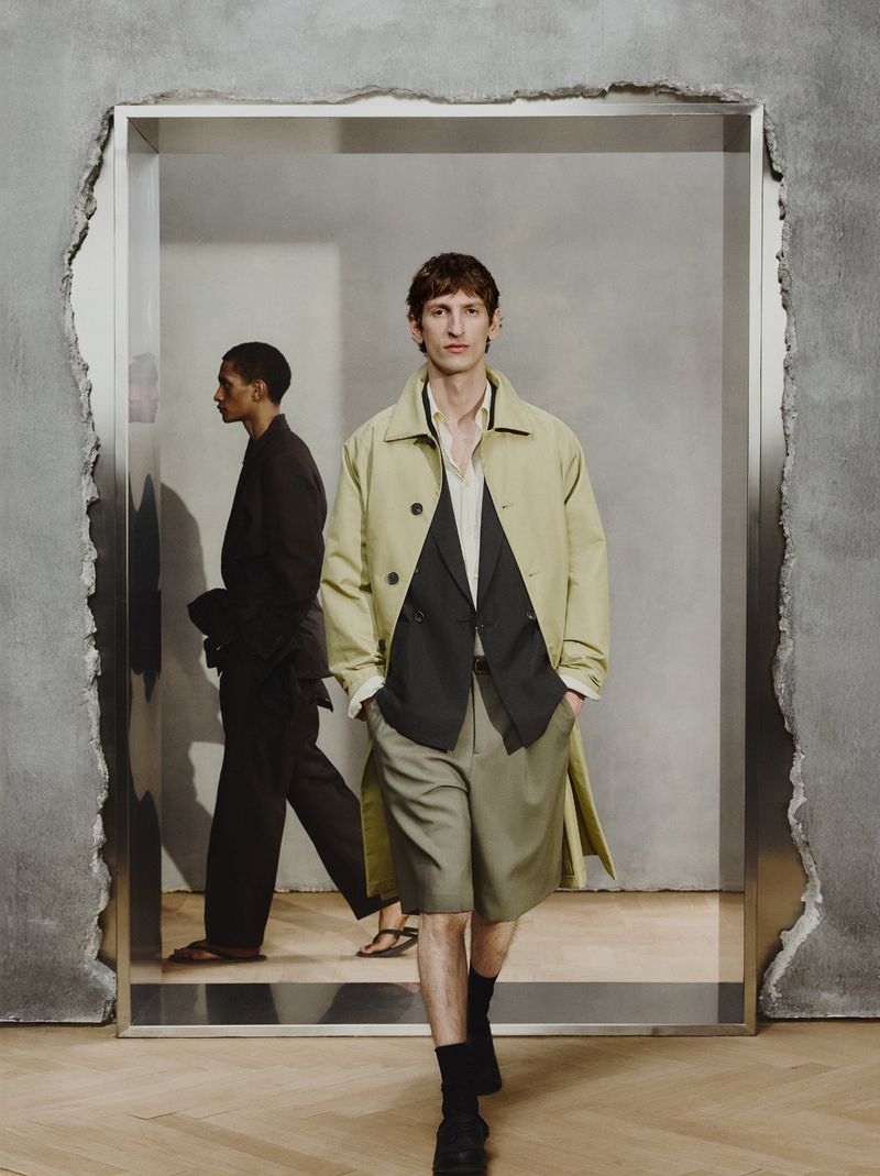 Two male fashion models pose indoors. One walks toward the camera, and the other walks away from the camera in the background.