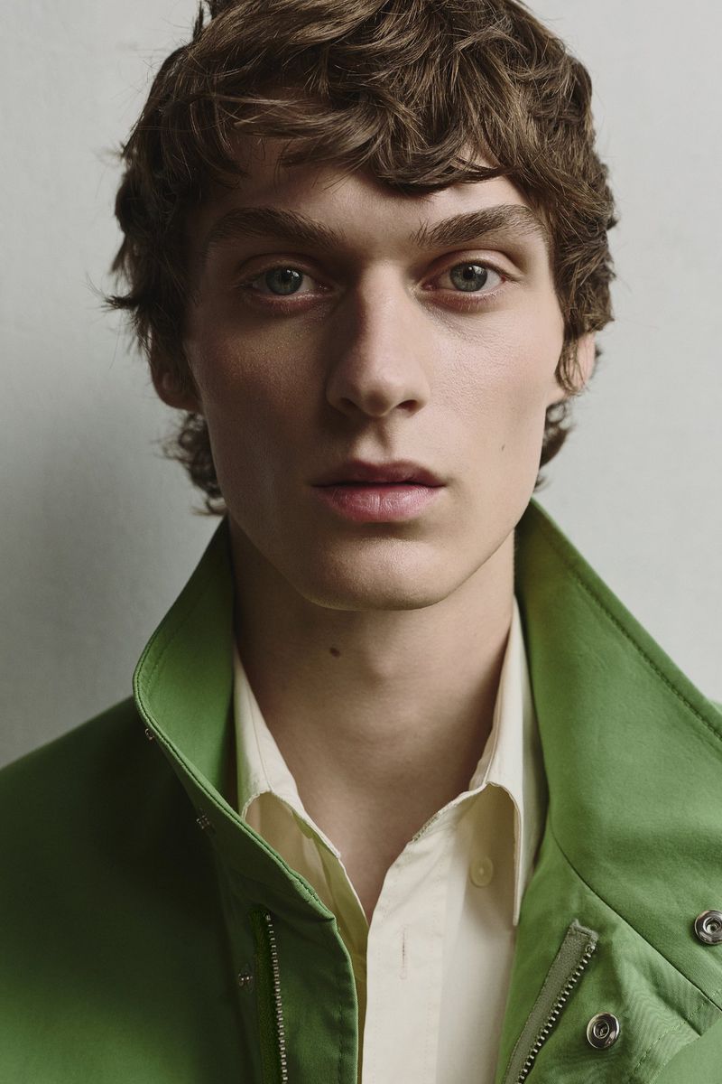 Close-up of a young adult male with curly brown hair, wearing a green jacket and light yellow collared shirt, against a plain background.