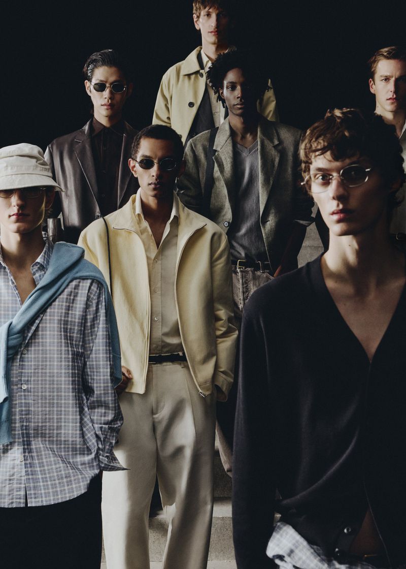 Group of male models in various casual and semi-formal outfits posing against a dark background for a fashion editorial shot.
