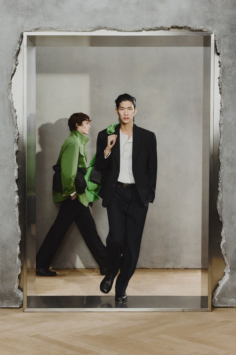 Two fashion models pose in a geometric frame wearing a black suit, white shirt and green jacket in a grey room with wood floors.