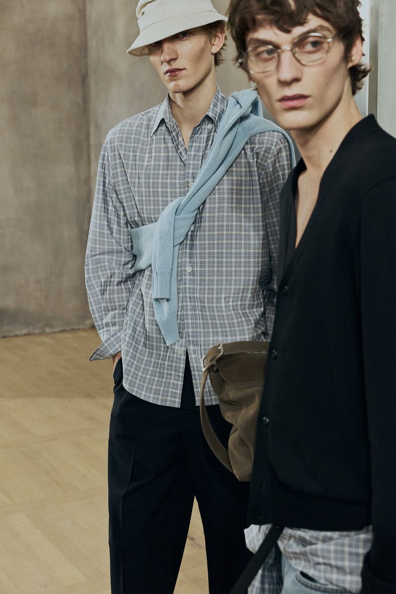 Two adult male fashion models in a studio setting, wearing casual shirts and pants; one wears glasses and a light-colored hat.