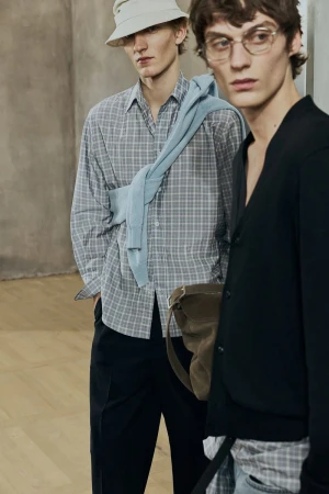 Two adult male fashion models in a studio setting, wearing casual shirts and pants; one wears glasses and a light-colored hat.