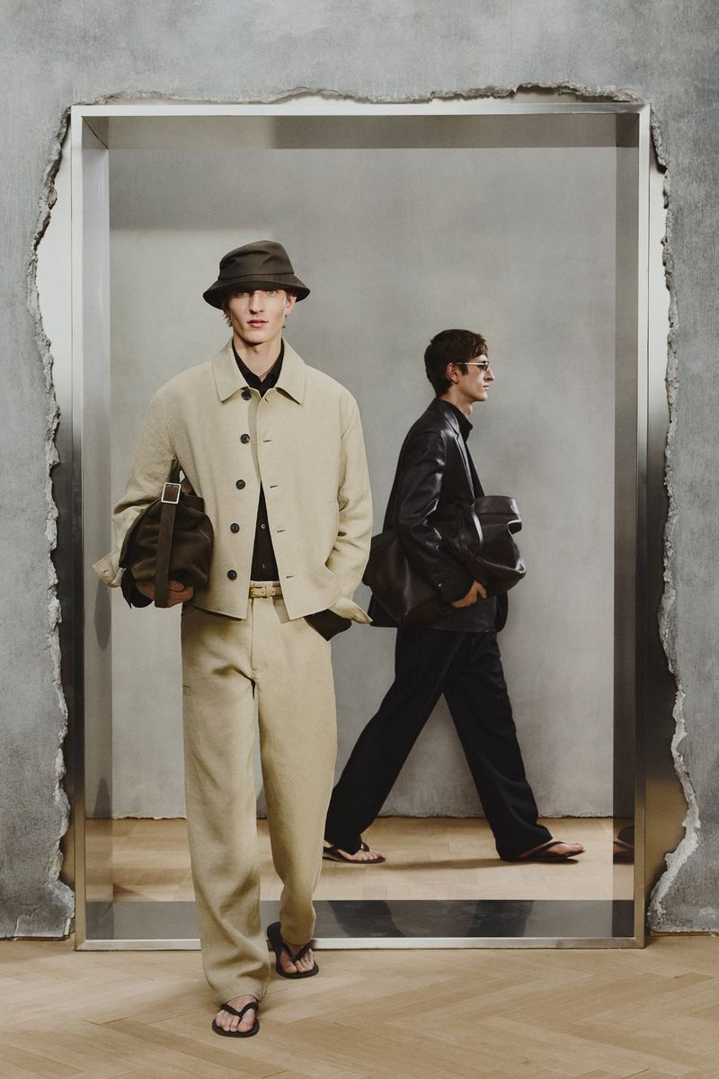 Two adult males in fashionable attire posing within a geometric doorway, showcasing a contemporary clothing collection with muted tones.