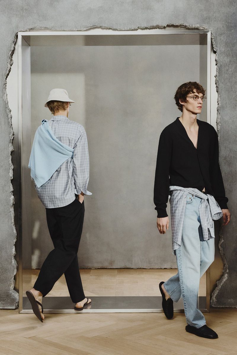 Two men wearing fashionable casual clothing, walking through a doorway with textured walls and a light-wood floor. 