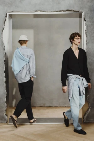Two men wearing fashionable casual clothing, walking through a doorway with textured walls and a light-wood floor. 