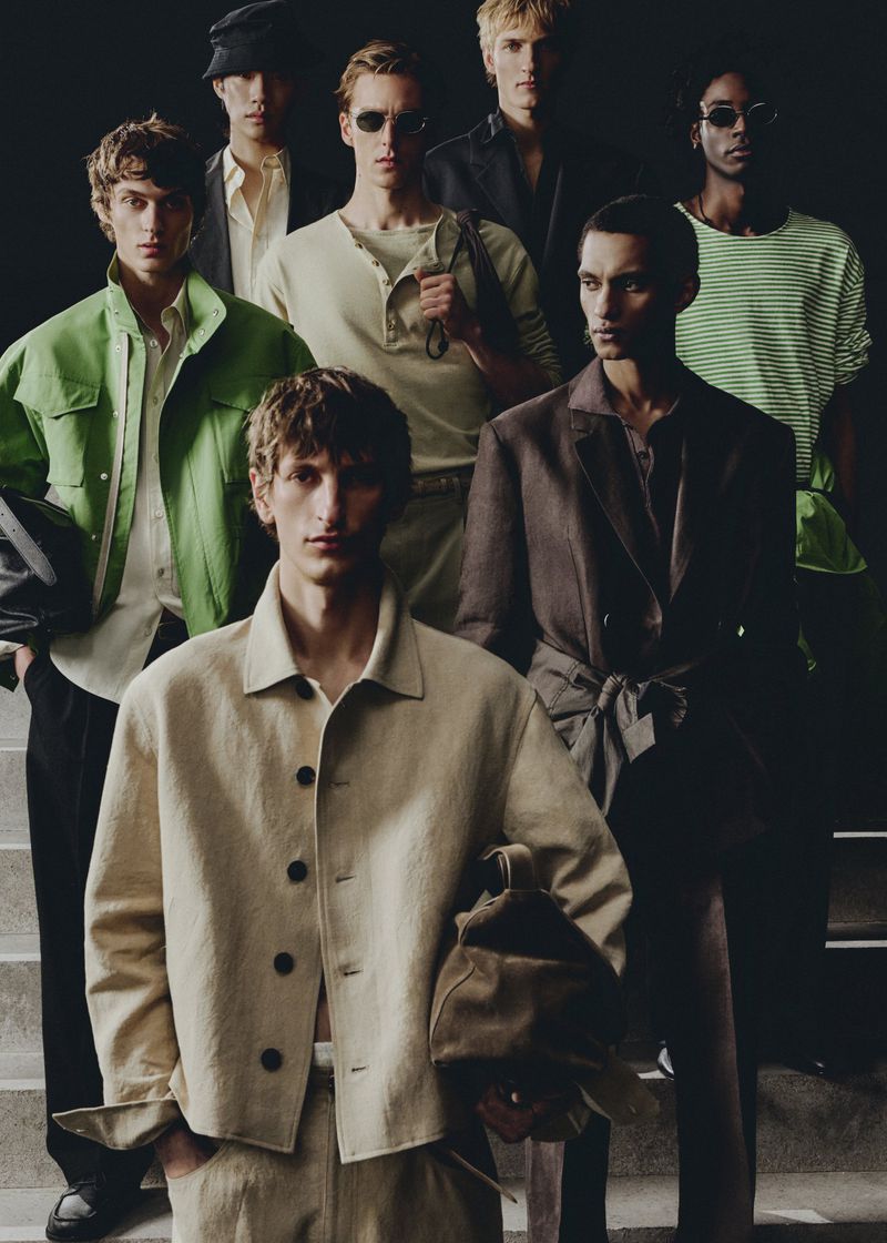 Group of male models wearing casual, neutral-toned clothing in a studio setting with dark background and bright directional lighting.