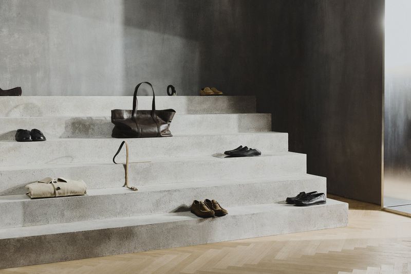 Arrangement of men's shoes, a leather tote bag, and a folded shirt on a concrete staircase, set against a neutral wall.