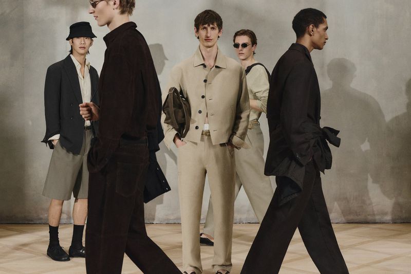 A group of men in stylish, neutral-toned outfits present a fashion collection against a neutral backdrop, captured in a medium shot.