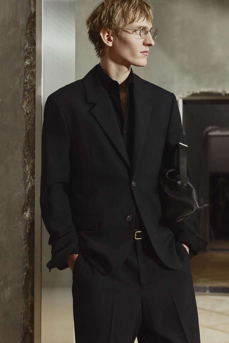 Blonde man in glasses, wearing a black suit with a leather bag, poses in a neutral setting with hands in pockets, looking to the side.