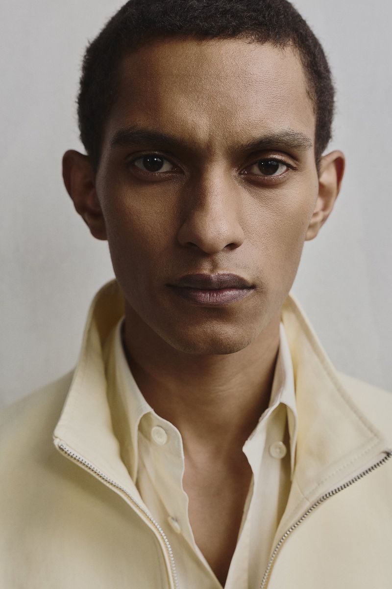 Portrait of a male model with dark hair and light skin, wearing an open white button-down shirt with a yellow jacket. Neutral background.