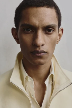 Portrait of a male model with dark hair and light skin, wearing an open white button-down shirt with a yellow jacket. Neutral background.